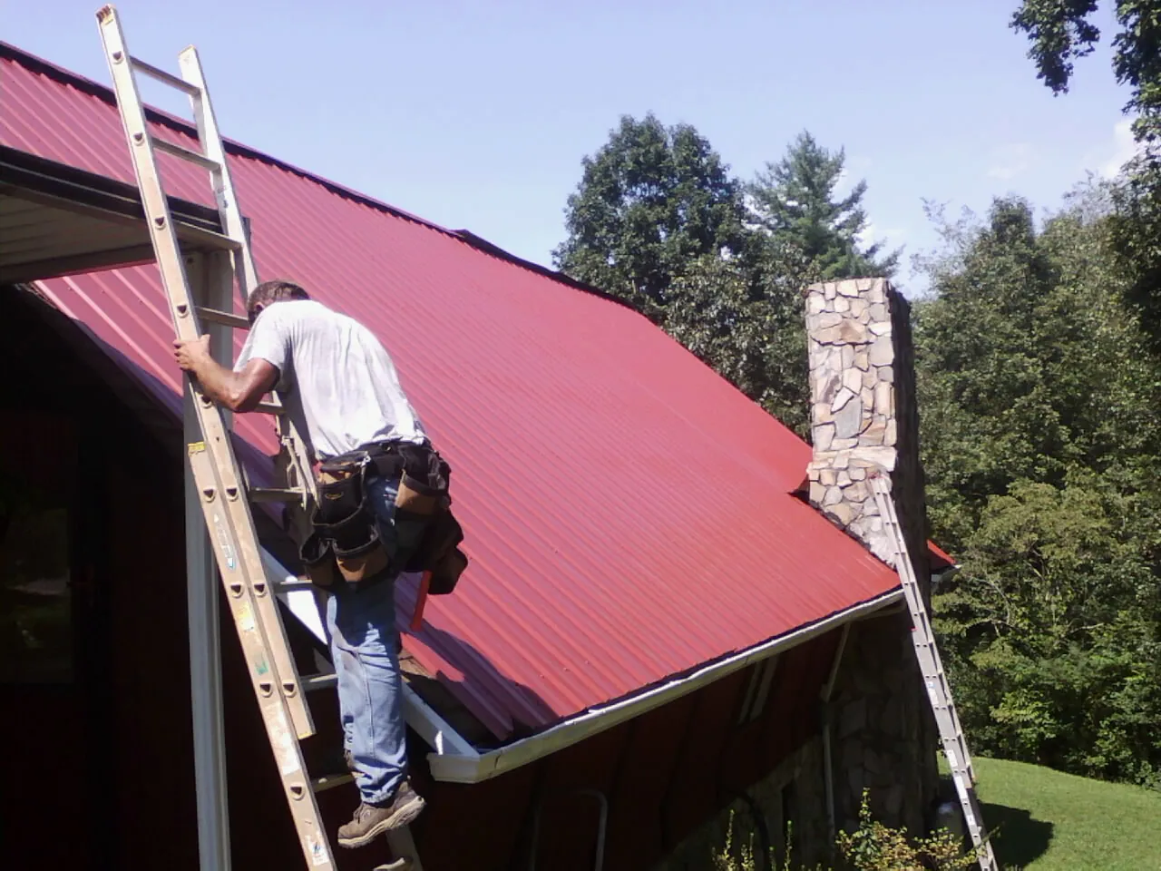 Roofer on a ladder beside a red metal roof and stone chimney — Soffit & Fascia Repair by Roofing Brothers in Conneaut